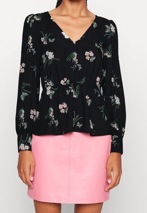 Kobieta ubrana w czarną floralną bluzkę z długim rękawem z marszczonym stanem oraz różową skórzaną mini spódnicę.