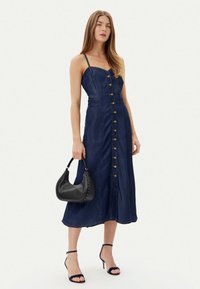 Abito midi in denim con corpino aderente, spalline regolabili, chiusura con bottoni frontale e gonna fluida. Abbinato a una borsa nera.