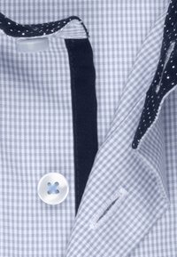 Primo piano del colletto di una camicia a quadretti piccoli azzurro chiaro e bianco con un bottone bianco e una fodera interna blu scuro con pois bianchi.