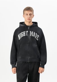 Young man wearing a black zip-up hoodie with "NIGHTMARE" in white letters and matching black pants, standing against a white background.