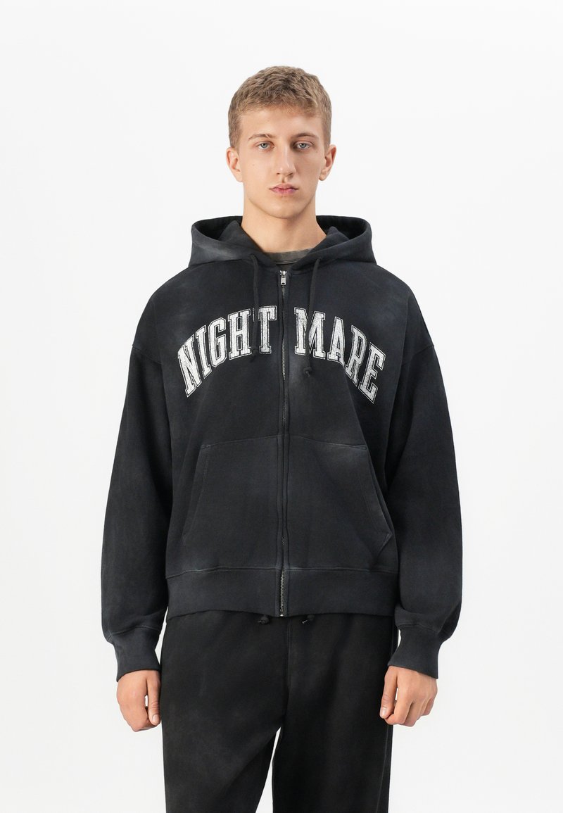 Young man wearing a black zip-up hoodie with "NIGHTMARE" in white letters and matching black pants, standing against a white background.