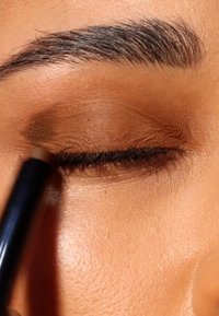 Brown eyeliner pencil applied to a closed eyelid. Skin tone is light brown with visible texture. Upper lashes are defined and dark.
