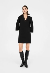 Robe noire à manches longues, col en V, avec des détails en dentelle aux poignets et au col. Portée avec des bottes noires montantes jusqu'aux genoux.