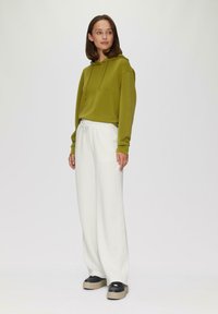 Zielona bluza z kapturem i sznurkiem, w połączeniu z białymi spodniami o szerokich nogawkach. Ubranie ma luźny styl i czarne buty na platformie z beżową podeszwą.