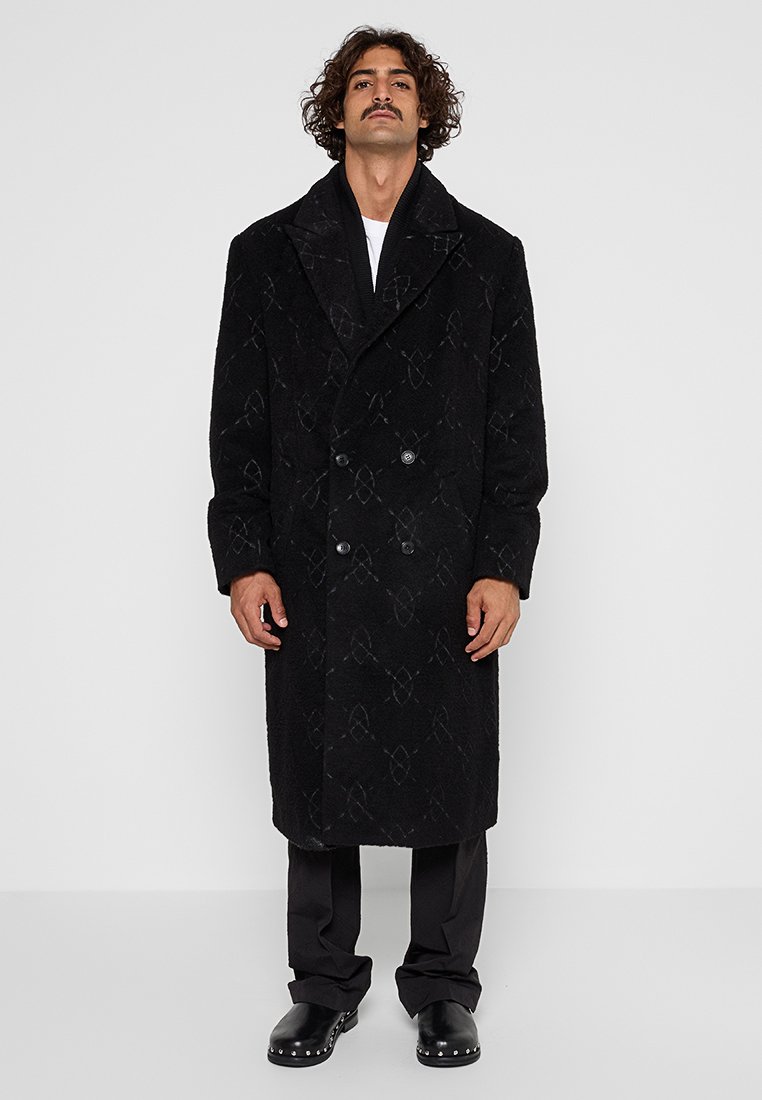 Manteau noir croisé avec une finition texturée et un motif subtil. Dispose d'un large col et de poches latérales. Porté avec un pantalon et des chaussures sombres.