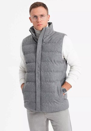 Gilet gris rembourré avec un col haut, fabriqué en tissu doux. Il présente une fermeture frontale et des poches latérales, porté sur un pull à col roulé blanc.