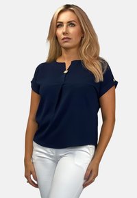Granatowa bluzka z krótkim rękawem i okrągłym dekoltem, z guzikiem przy kołnierzyku i luźnym krojem. Noszona z białymi spodniami.