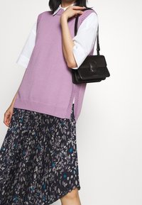 Vaaleanlila neuleliivi, jossa on nyöritykset, käytössä valkoisen paidan päällä. Asusteena musta nahkakäsilaukku ja kuviollinen taittoskirt.