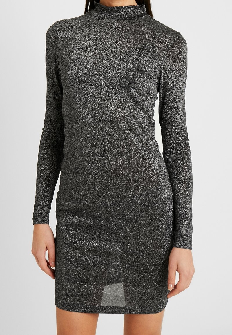 Robe ajustée manches longues scintillante gris foncé avec col montant, portée par une personne debout sur un fond clair uni.