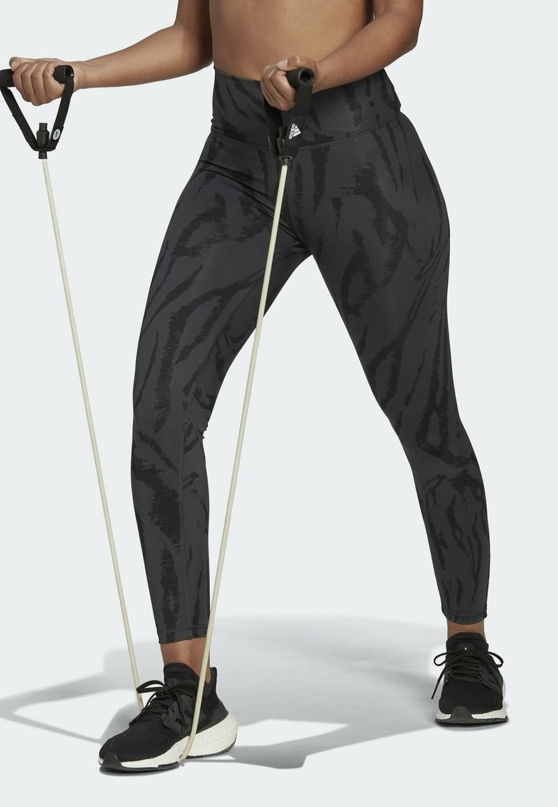 Zwarte legging met een subtiel tijgerstreepje, hoge taille, en rekbaar, vochtregulerend materiaal; gecombineerd met zwarte sportschoenen.