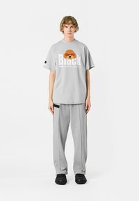 Camiseta de gran tamaño en gris claro con un gráfico de un perro marrón texturizado y texto en negro en negrita. Combinada con pantalones gris claros y zapatos negros texturizados.