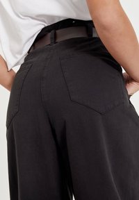 Czarne spodnie o szerokich nogawkach z teksturowanej tkaniny, z dwoma tylnymi kieszeniami i pasem. Stroje zawierają białą koszulę widoczną u góry.