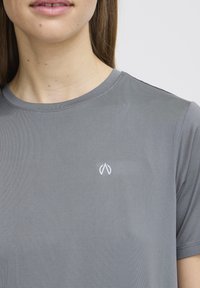 Pilkos spalvos trumpomis rankovėmis marškinėliai, pagaminti iš lygaus audinio, su mažu baltu logotipu kairėje krūtinės pusėje ir apvaliu iškirpimu.