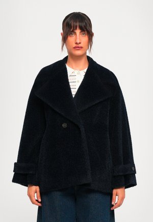 Woman with dark hair and bangs wearing a black textured oversized coat over a white patterned shirt and dark blue jeans against a plain background.