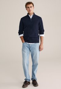 Mornarsko plavi pulover s patentnim zatvaračem na ovratniku, uparen s bijelom košuljom s ovratnikom. Svijetloplave traperice i smeđe cipele upotpunjuju odjevnu kombinaciju. Dizajn tkanine s teksturom.