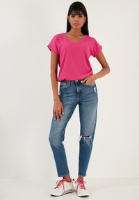 Pink, short-sleeve, V-neck top paired with distressed blue jeans. Model wears white sneakers with red laces. Simple accessories and a neutral backdrop.
