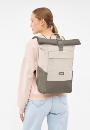 Woman wearing a beige and olive roll-top backpack, dressed in pale pink sweatshirt and light blue jeans, shown from the back and side.