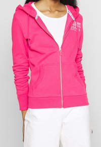 Felpa con zip di colore rosa acceso in pile, dotata di tasca a marsupio, polsini e bordo a costine, con un logo bianco sul petto.