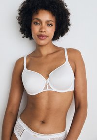 White bra with smooth cups, adjustable straps, and a mesh center panel. Features a gold-tone accent at the front.