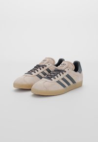 Beige tenisky Adidas Gazelle s černými pruhy, černými tkaničkami a gumovými podrážkami, zobrazené z předního úhlového pohledu na bílém pozadí.