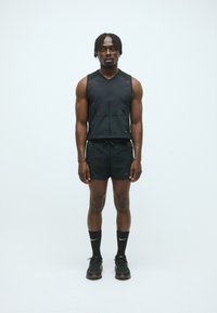 Athletischer Mann steht, trägt ein schwarzes ärmelloses Zip-Top, schwarze Shorts, schwarze Nike-Socken und schwarze Laufschuhe vor einem schlichten weißen Hintergrund.