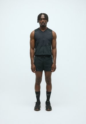 Athletischer Mann steht, trägt ein schwarzes ärmelloses Zip-Top, schwarze Shorts, schwarze Nike-Socken und schwarze Laufschuhe vor einem schlichten weißen Hintergrund.