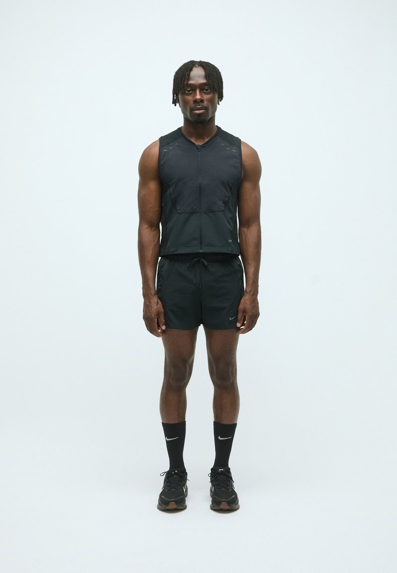 Athletischer Mann steht, trägt ein schwarzes ärmelloses Zip-Top, schwarze Shorts, schwarze Nike-Socken und schwarze Laufschuhe vor einem schlichten weißen Hintergrund.