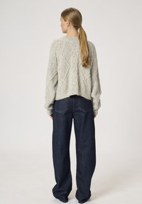 Hellegrau gestrickter Pullover mit Zopfmuster, überschnittenen Schultern und gerippten Bündchen, kombiniert mit weit geschnittenen dunkelblauen Jeans.