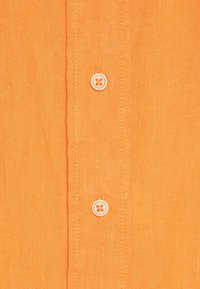 Camicia in lino arancione a maniche corte con chiusura frontale a bottoni bianchi e un tessuto testurizzato. I semplici dettagli delle cuciture aggiungono una sottile definizione.