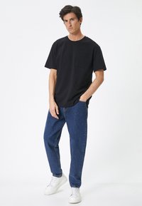 Camiseta de algodón negra con mangas cortas y un bolsillo en el pecho, combinada con jeans de mezclilla azul claro y zapatillas blancas sobre un fondo sencillo.