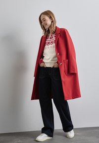 Roter, knielanger Mantel mit strukturiertem Design, zwei Vordertaschen und großen Knöpfen. Cremerfarbener Pullover mit rosa gemusterten Akzenten. Dunkelblaue Jeans und weiße Sneakers.