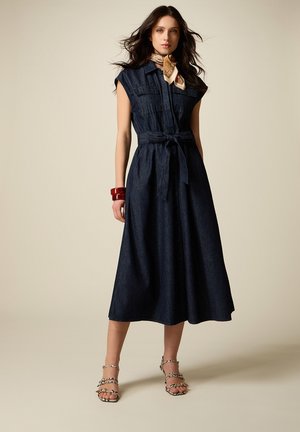 Kvinde iført mørkeblå midi-kjole uden ærmer med bælte, dyreprintede sandaler med hæl, røde armbånd og et mønstret halstørklæde, stående foran beige baggrund.