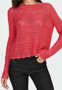 Koraalroze gebreide trui met een open gehaakt patroon, geribde halslijn en een gerimpelde zoom. Gecombineerd met donkere spijkerbroek.