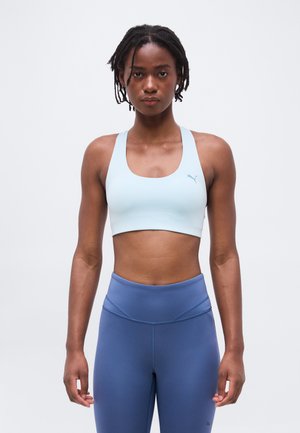 Fiatal nő fonott hajjal, világoskék sportmelltartót és magas derekú kék leggingset visel, egyszínű fehér háttér előtt áll.