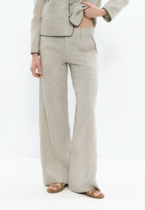 Personne portant un pantalon large à rayures beige et blanc, un blazer assorti et des sandales ouvertes marron, debout devant un fond blanc.