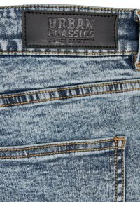 Jean en denim avec un délavage bleu, des surpiqûres orange en contraste et une étiquette en cuir noir embossée avec "URBAN CLASSICS DENIM FACTORY."