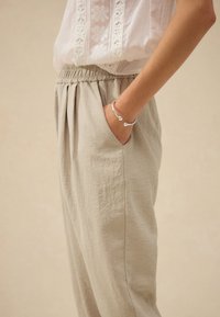 Heledad beežid püksid tekstuurse viimistlusega, millel on elastne vöökoht ja küljetaskud. Randmel on hõbedane käevõru.