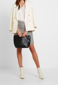 Sac à main en cuir noir à texture lisse ; porté avec un blazer crème croisé et une jupe à carreaux, complétés par des bottines beiges.