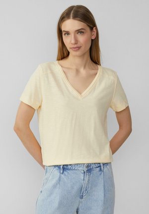 T-shirt à manches courtes et col en V en tissu doux de couleur jaune clair. Coupe décontractée avec un minimum de détails de couture et une silhouette relaxée, associée à un jean bleu clair.