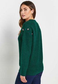 Emeraldgroene gebreide trui met een textuurafwerking, afgewerkt met zilveren knopen op de schouders en geribbelde manchetten. Lange mouwen en een relaxte pasvorm.