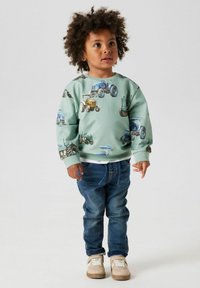 Felpa verde menta con stampa di trattore, abbinata a jeans blu e sneaker beige. L'outfit è casual e comodo.