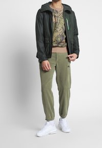 Giacca nera con cappuccio, felpa con motivo camouflage, pantaloni jogger verde oliva e sneakers bianche. Presenta cerniere e polsini elasticizzati.