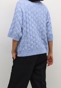 Dik lichtblauw gebreide trui met korte mouwen, voorzien van een gestructureerd kabelpatroon en geribde zoom, gecombineerd met een zwarte broek.