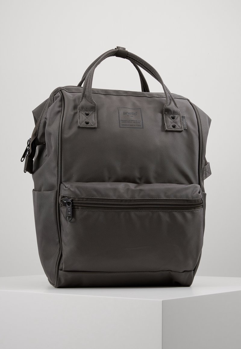Šedý nylonový batoh s dvojitými horními uchy, přední kapsou na zip, texturovaným povrchem a nenápadnou značkovou nálepkou. Čtvercový tvar s vyztuženým švem.