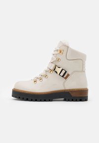 Bottes chevilles en cuir blanc avec quincaillerie dorée, doublure en peluche et semelle noire texturée. Présente une large sangle avec un accent de boucle.
