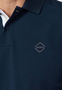 Primo piano di una polo blu navy con colletto nero e una piccola toppa con il logo Hackett sul petto.