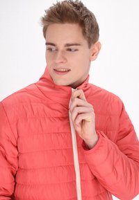 Koralļu krāsas siltās jakas ar rāvējslēdzēju un stāvošu apkakli, gluda tekstūra, izlīdzinoša raksts un krēmkrāsas rāvējslēdzēja detaļa.
