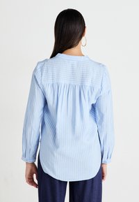 Blusa a rayas azul claro con mangas largas, dobladillo curvado y detalles de espalda fruncida. El tejido parece suave con una textura ligera.