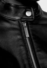 Zbliżenie na kołnierz czarnej skórzanej kurtki z metalowym zatrzaskiem i widocznym suwakiem z wygrawerowanym napisem "MK".