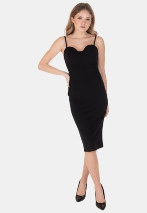 Donna in piedi con una mano sul fianco, indossa un abito midi nero aderente con sottili spalline e tacchi alti neri su sfondo bianco.